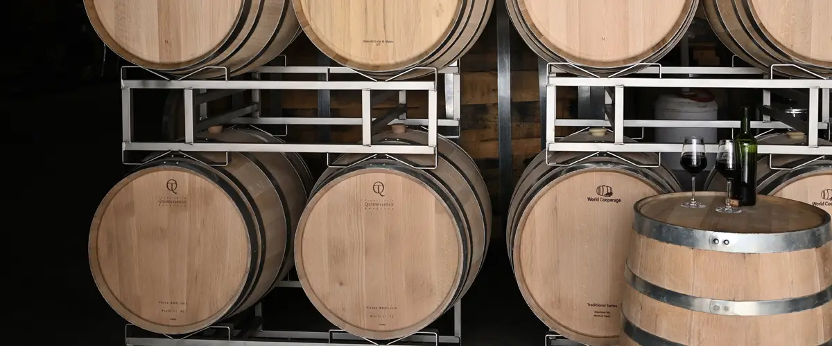 Partner-Küferei Viele Fässer von verschiedenen Herstellern sind auf dem Bild liegend auf Metallfasslagern zu erkennen. Unten rechts steht ein Fass an der Seite mit zwei gefüllten Rotweingläsern und einer Flasche.