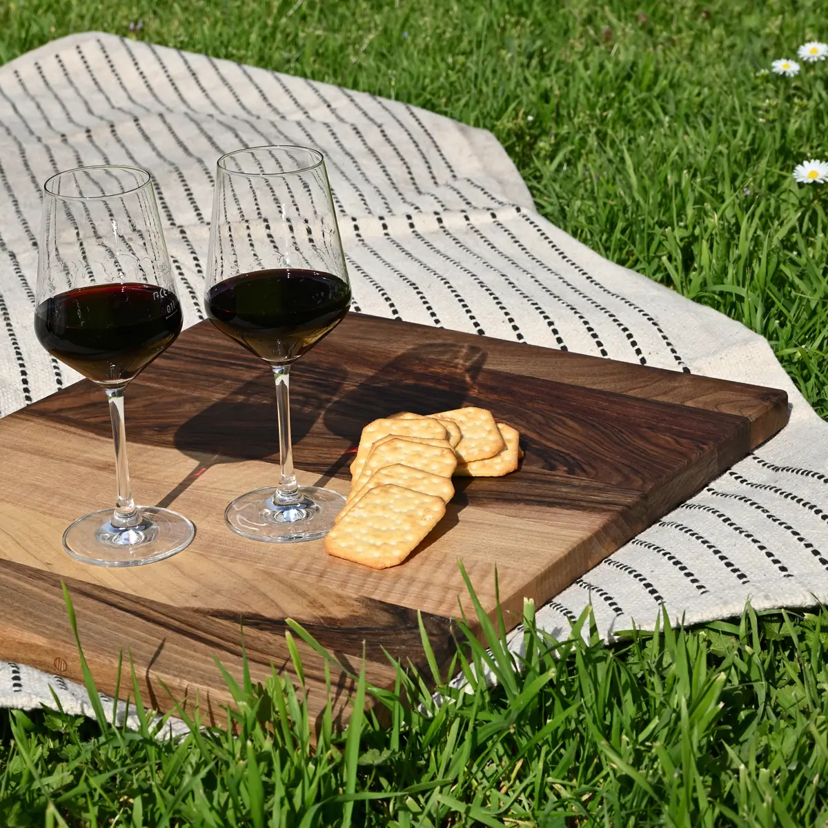 Küche und Fass Auf einer Wiese liegt eine Picknickdecke mit einem Walnussbaumholzbrett, Kräckern und Weingläser mit Rotwein.